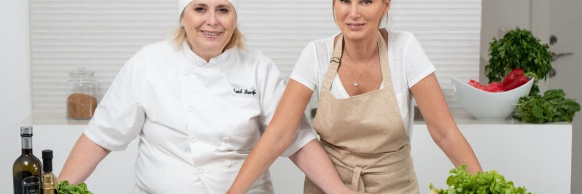 Two female chefs with variety of fresh ingredients in a kitchen setting, showcasing culinary preparation.
