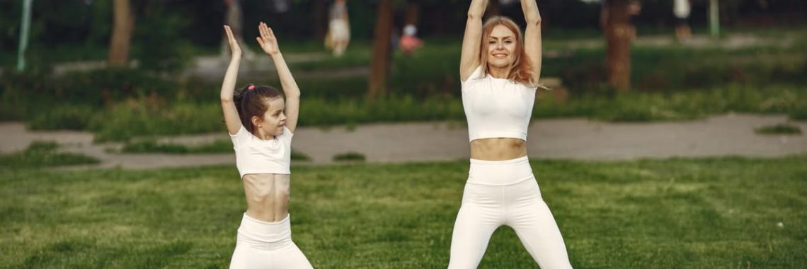 Mother and daughter in matching activewear doing exercises outdoors on a sunny day.